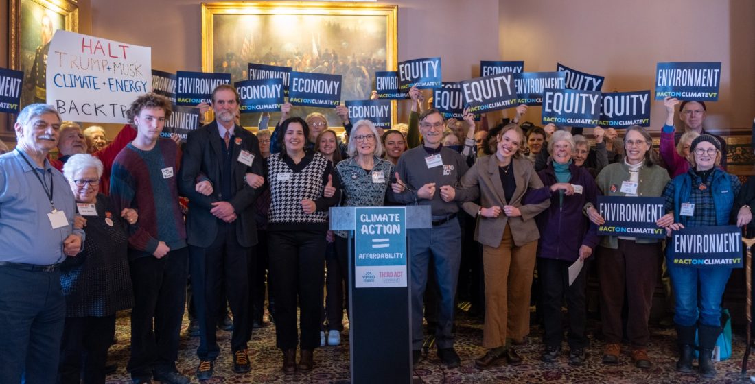 Group of people with linked arms and signs behind a podium in the Vermont statehouse.